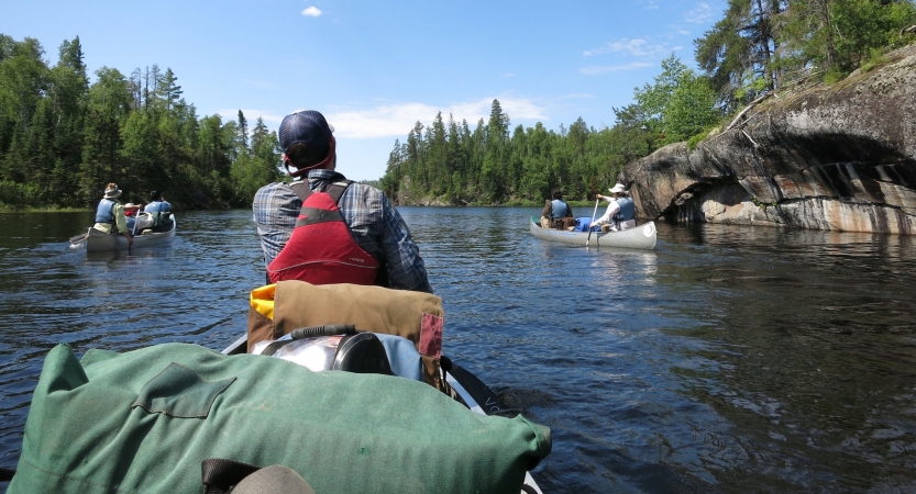 From the back of a canoe, the person in front navigates, and two other manned canoes are pictured in the distance. 